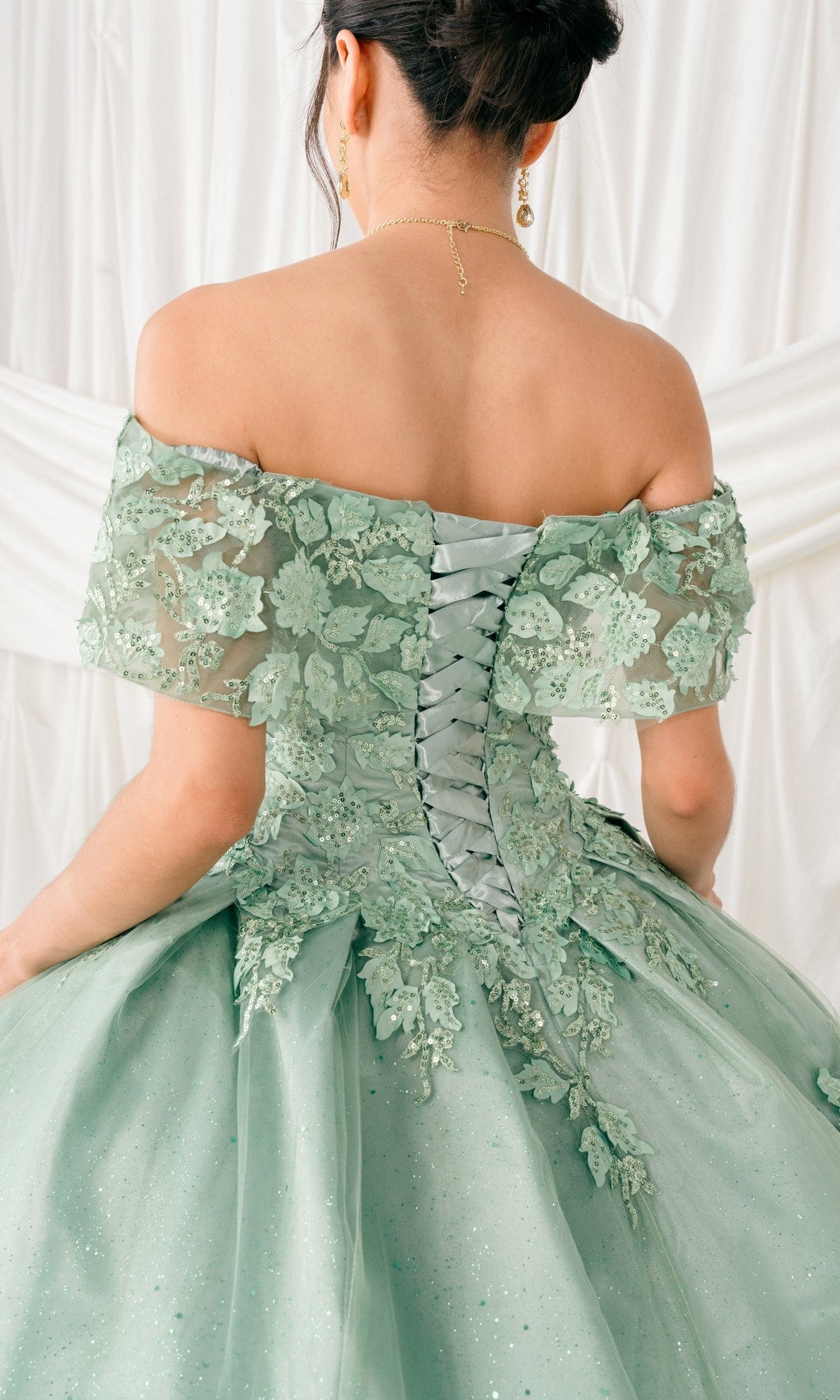 A woman in the Quinceanera Ball Gown 8045J, featuring off-the-shoulder design, 3D floral details, and glitter lace embroidery, stands with her back to the camera against a white draped background.
